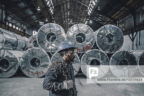 Seitenansicht eines männlichen Arbeiters  der eine Kette trägt  während er an aufgerollten Stahlblechen in der Fabrik steht