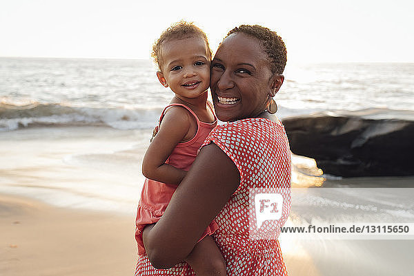 Portrait of mother carrying daughter while standing at beach during sunset