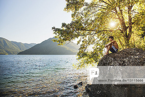 Nachdenklicher Teenager ruht sich auf einem Felsen aus und schaut an einem sonnigen Tag auf den See