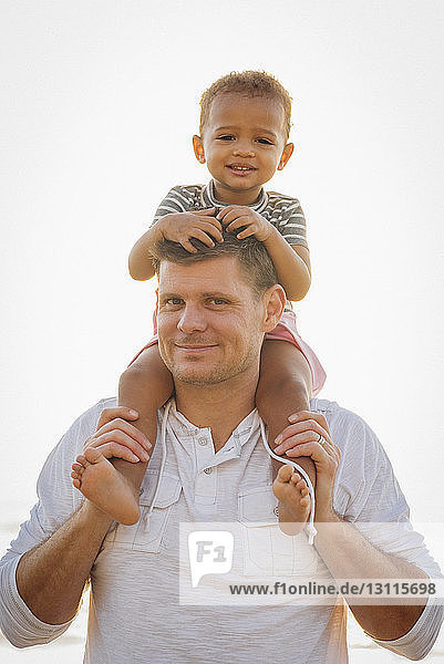 Porträt eines Vaters  der seine Tochter auf den Schultern trägt  während er am Strand vor klarem Himmel steht