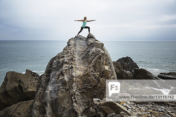 Frau übt die Worrier 2-Pose auf Felsen am Meer