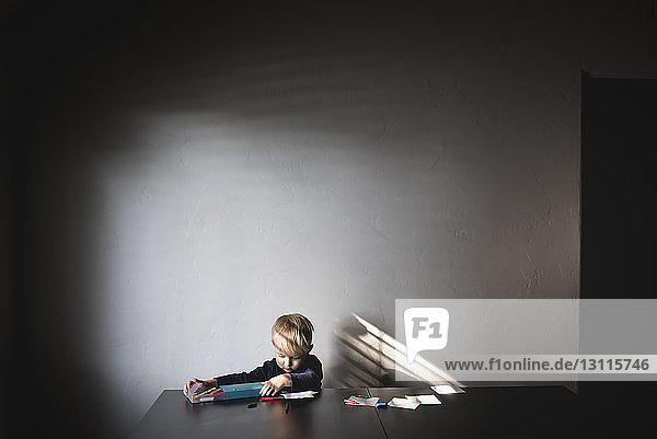 Junge entfernt Buntstifte aus der Schachtel  während er am Tisch an der Wand sitzt