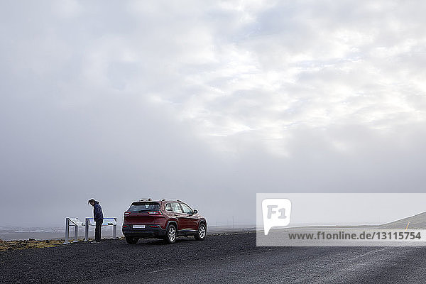 Side view of man standing by car on road against cloudy sky