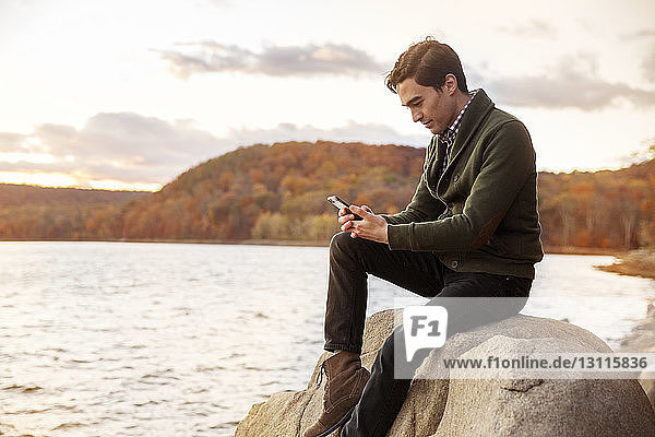 Mann benutzt Mobiltelefon  während er auf einem Felsen am Seeufer sitzt