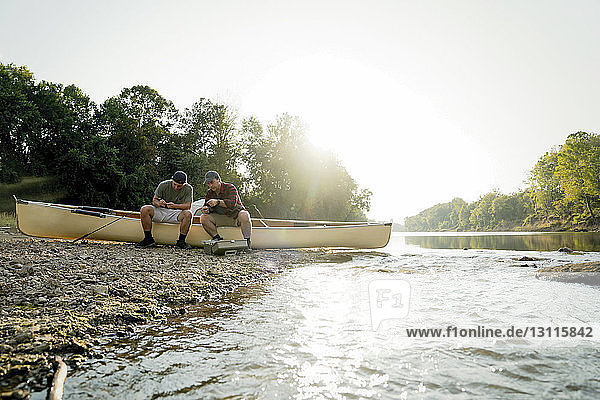 Männliche Freunde passen Angelgeräte an  während sie am Seeufer bei klarem Himmel auf einem Boot sitzen
