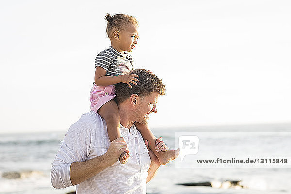Vater trägt Tochter auf den Schultern  während er am Strand vor klarem Himmel steht