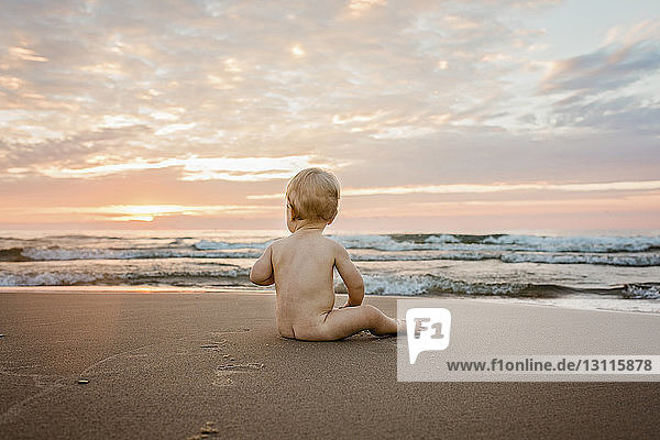 Nackter Junge in voller Länge  der bei Sonnenuntergang auf Sand am Strand vor bewölktem Himmel sitzt