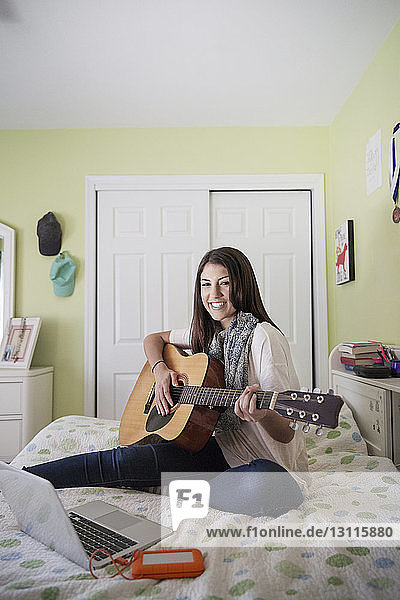 Porträt einer glücklichen Teenagerin  die Gitarre spielt  während sie zu Hause am Laptop sitzt