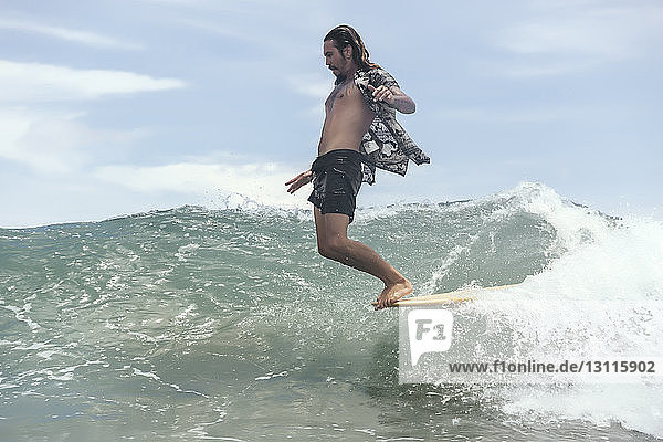 Man surfing while standing on surfboard's edge in sea against sky