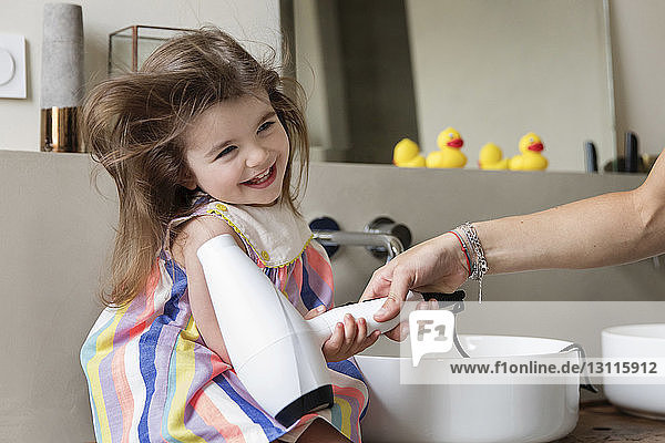 Abgehackte Hand einer Mutter  die das Haar ihrer glücklichen Tochter trocknet und zu Hause auf dem Waschbecken im Badezimmer sitzt
