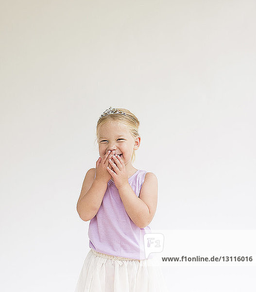 Cheerful shy girl wearing tiara covering mouth while standing against white background