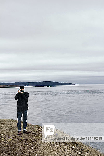 Mann fotografiert  während er am Ufer vor bewölktem Himmel steht