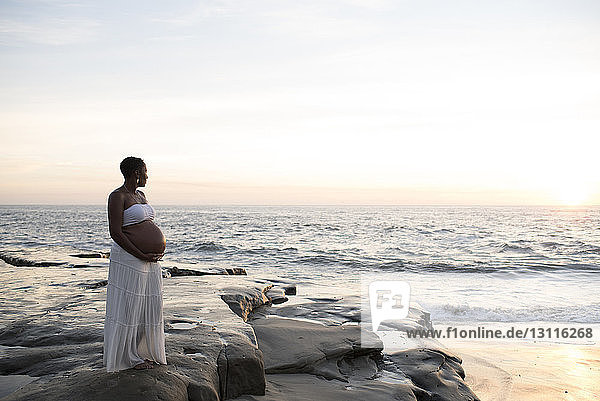 Pregnant woman standing at beach against sky during sunset