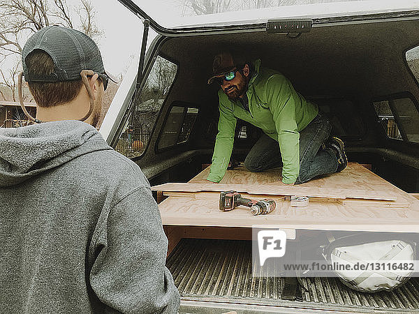 Man talking to male friend while kneeling on wood in vehicle trunk