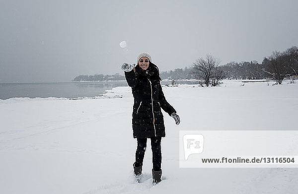 Porträt einer lächelnden Frau  die einen Schneeball wirft  während sie am schneebedeckten Strand gegen den Himmel steht