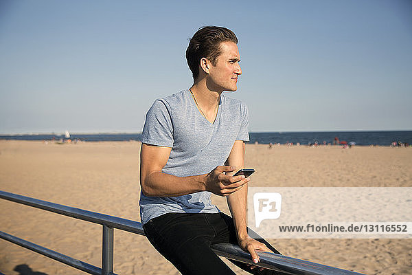 Junger Mann sitzt am Strand auf einem Geländer vor klarem Himmel