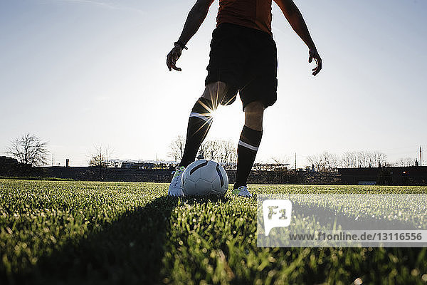 Niedriger Teil eines Mannes  der bei Sonnenuntergang auf einem Rasenplatz vor klarem Himmel Fußball spielt