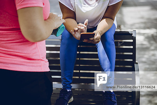 Low section of woman using smart phone while sitting on bench with friend standing in foreground