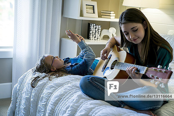 Girl using smart phone while sister playing guitar on bed