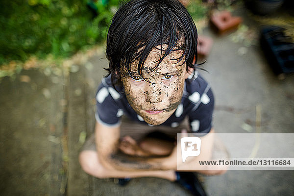 High angle portrait boy with dirty face sitting on footpath at yard during rainy season