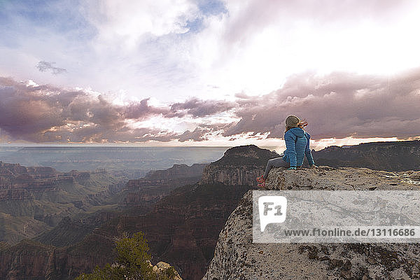 Rückansicht einer Frau  die Berge betrachtet  während sie auf einem Felsen vor bewölktem Himmel sitzt