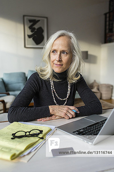 Thoughtful senior woman looking away while sitting by laptop computer on table at home