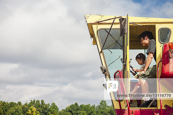 Side view of father and daughter sitting in tractor at farm against cloudy sky