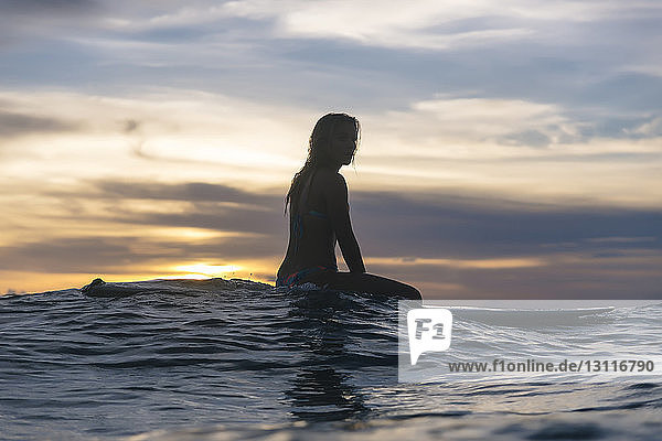 Side view of young woman sitting on surfboard in sea against cloudy sky during sunset