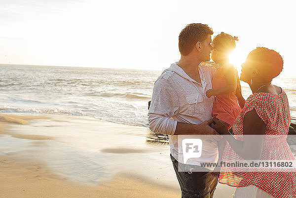 Glückliche Eltern spielen mit ihrer Tochter  während sie bei Sonnenuntergang am Strand gegen den Himmel stehen