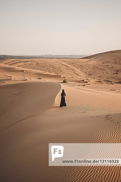 Rear view of woman walking at Sahara Desert against clear sky during sunset