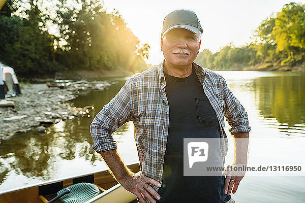 Portrait of senior man with hands on hip standing by boat at lakeshore during sunset