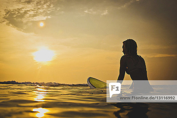 Woman sitting on surfboard in sea during sunset