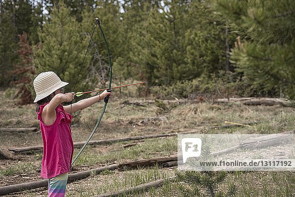 Side view of girl aiming with bow and arrow while standing against trees in forest