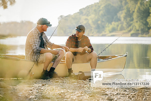 Friends talking while sitting by fishing rods on boat at lakeshore