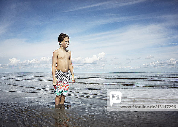 Nachdenklicher Junge ohne Hemd steht im Meer gegen den Himmel