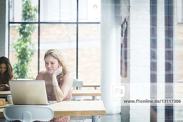 Frau mit Hand am Kinn  die einen Laptop benutzt  während sie mit einem Freund im Hintergrund durch ein Fenster in einem Café sitzt