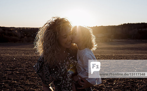 Liebende Tochter küsst fröhliche Mutter  während sie von ihr bei Sonnenuntergang auf dem Feld getragen wird