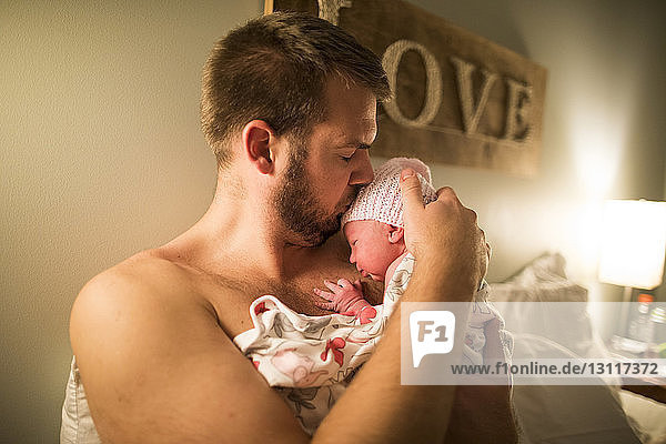 Shirtless father kissing newborn daughter in bedroom at home