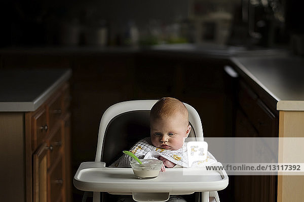 Kleiner Junge betrachtet Essen in Schüssel  während er auf einem Hochstuhl sitzt