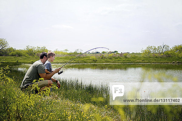 Side view of father and son fishing in pond against sky