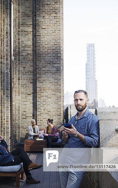 Porträt eines Mannes  der ein Mobiltelefon benutzt  während seine Kollegen im Hintergrund sitzen