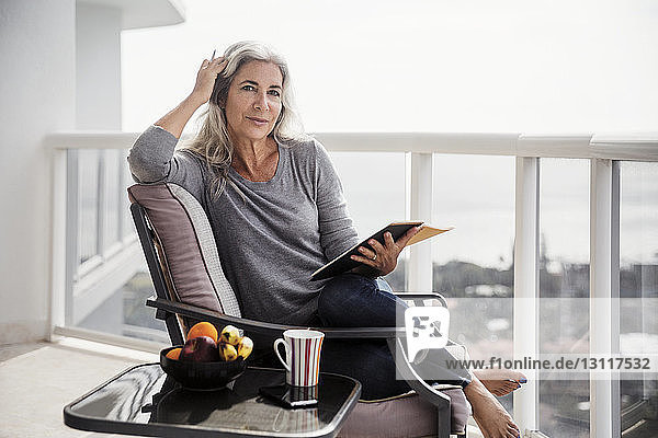 Porträt einer selbstbewussten Frau  die ein Buch hält  während sie auf einem Stuhl auf dem Balkon sitzt
