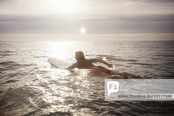 Rückansicht einer Surferin  die bei Sonnenuntergang auf einem Surfbrett im Meer liegt