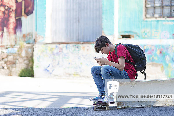 Seitenansicht eines Schülers mit Skateboard  der ein Mobiltelefon benutzt  während er in der Stadt sitzt