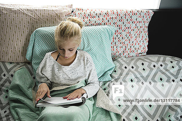 Hochwinkelansicht eines Mädchens  das ein Buch liest  während sie sich im Schlafzimmer auf Kissen stützt