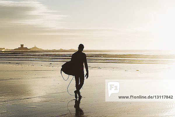 Silhouetten-Mann mit Surfbrett beim Strandspaziergang bei Sonnenuntergang