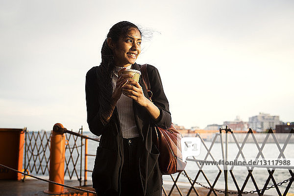 Glückliche Frau hält Einweg-Kaffeetasse  während sie am Fluss steht