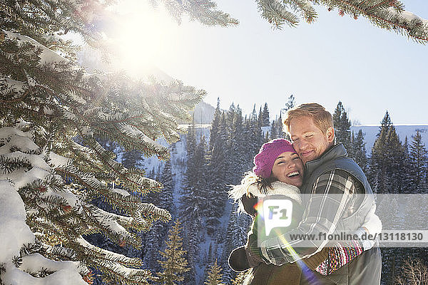 Couple embracing while standing in snowy forest against clear sky
