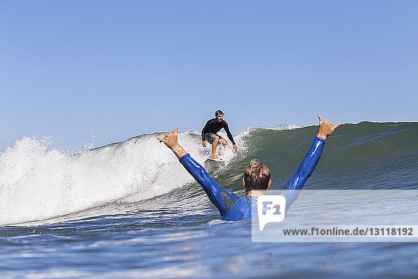 Mann jubelt  während er einen Freund beim Surfen auf dem Meer vor klarem blauen Himmel betrachtet