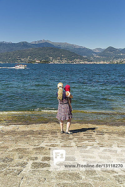 Rear view of mother carrying daughter while standing by lake against clear blue sky during sunny day